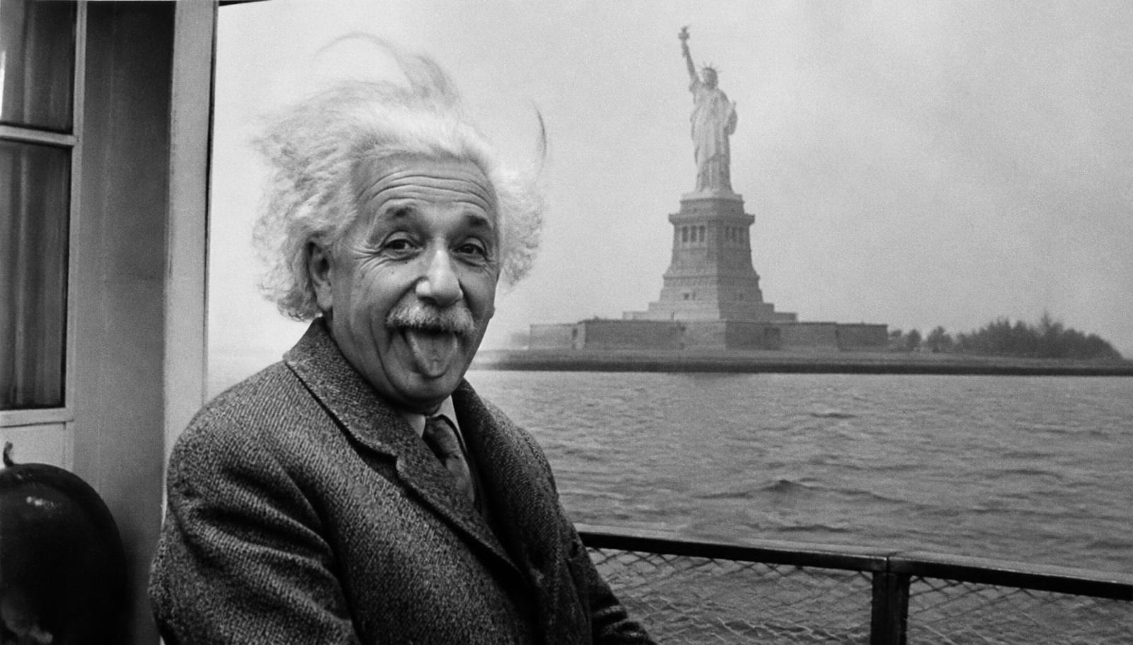 Black and white photograph of a white-haired scientist sticking his tongue out, with the Statue of Liberty behind him